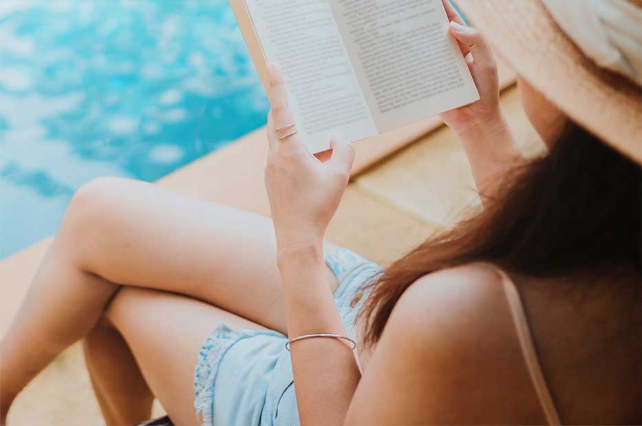 Lady reading a book and sitting next to a swimming pool.
